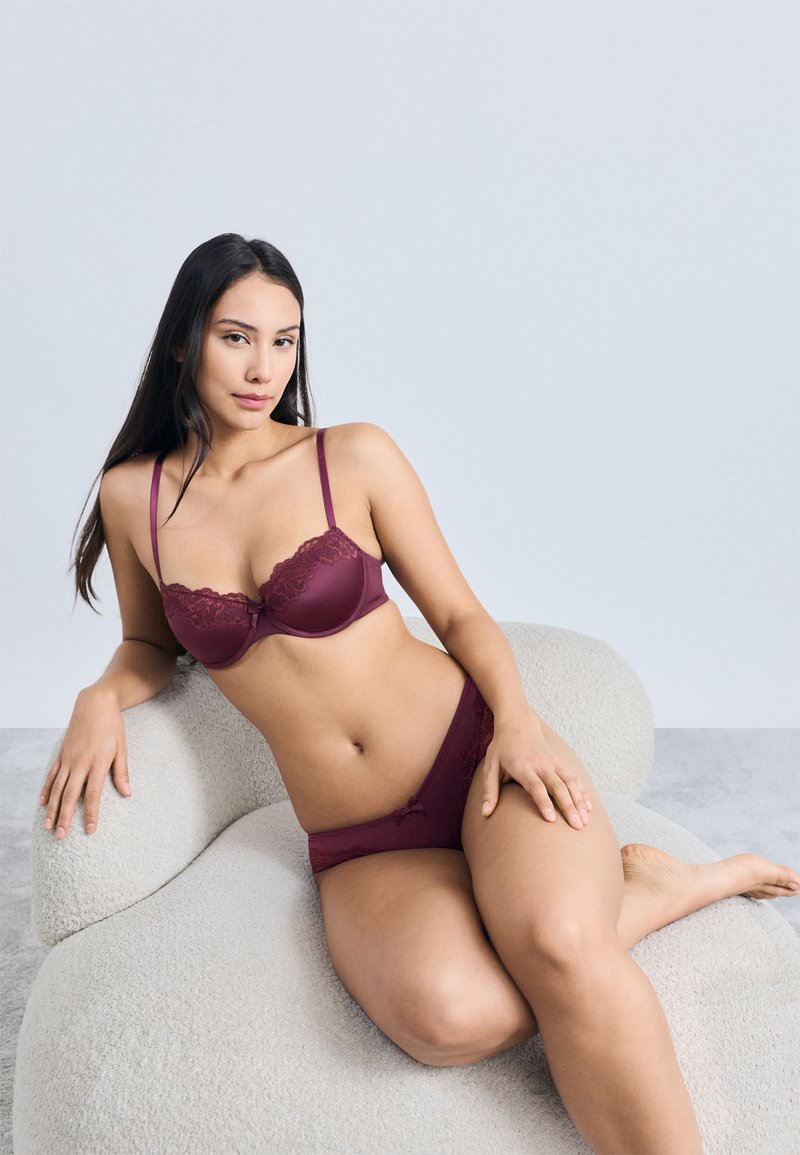 Vīnogu krāsas apakšveļas komplekts ar mežģīņu maliņu krūšturī ar veidnēm un atbilstošām biksītēm, abas izgatavotas no gludas auduma, attēlotas uz neitrāla fona.