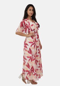 Robe maxi à imprimé floral avec des tons de rose et de pêche. Présente une taille smockée et de courtes manches à volants. Portée avec des talons blocs transparents.