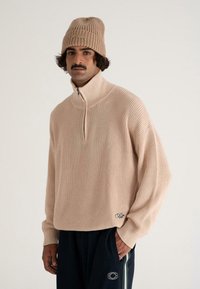 Mann mit Schnurrbart, der eine beigefarbene Strickmütze, einen passenden beigen Halbzip-Pullover und schwarze Hosen mit gestreiften Details und Logos trägt.