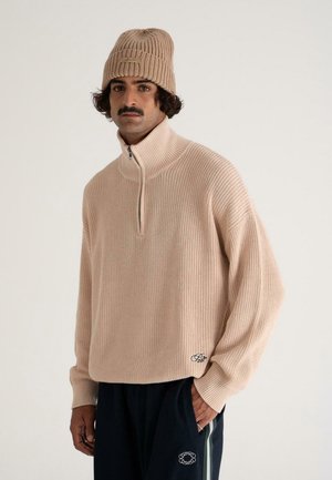 Man with mustache wearing a beige knit beanie, matching beige half-zip sweater, and black pants with striped detailing and logos.