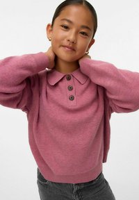 Ung tjej med mörkt hår i en rosa tröja med knappar, som håller om sin nacke och leende mjukt mot en helt vit bakgrund.