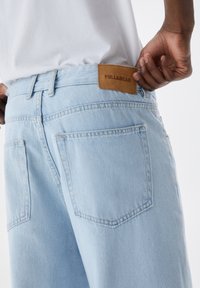 Pantalones vaqueros de mezclilla azul claro con corte relajado, que presentan un parche de logo de cuero marrón en la cintura y dos bolsillos traseros con detalles de costura.
