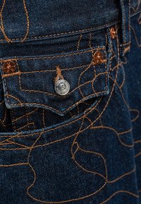 Tkanina dżinsowa w ciemnoniebieskim kolorze, z brązowym przeszyciem w falisty wzór oraz metalowymi guzikami na kieszeniach.