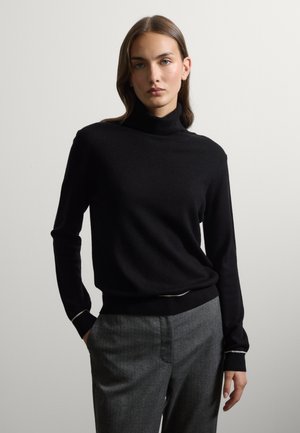Joven mujer con el pelo castaño liso, vistiendo un jersey negro de cuello alto y pantalones grises, de pie frente a un fondo claro liso.