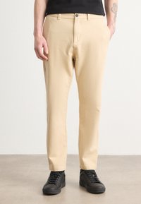 Pantalons en coton beige avec une coupe slim, dotés d'une fermeture à bouton, de poches latérales, et portés avec des sneakers noires.