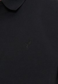 Μαύρο πόλο μπλουζάκι με μικρό κεντημένο λογότυπο κολιμπρί στο αριστερό στήθος, με διπλωμένο γιακά.