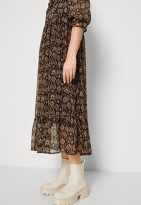Robe maxi noire à motifs avec un ourlet à volants, présentant des tons terreux et une silhouette fluide. Associée à des bottines crème à la cheville.