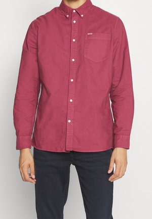 Mand iført en langærmet, knaplukket, mørkerød skjorte med brystlomme, kombineret med sorte bukser, mod en ensartet hvid baggrund.