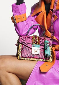 Femme portant une veste en cuir violette et orange, tenant un sac à main à motif peau de serpent multicolore avec des détails dorés et un pompon.