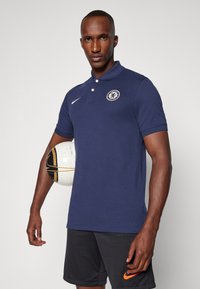 Donkerblauwe poloshirt met een kraag en korte mouwen, met een Chelsea FC-logo op de borst. Het model houdt een voetbal vast.