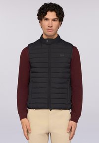 Černá vyp padded vesta s vysokým límcem, vertikálním prošíváním, předním zipem a logem. Nositá přes burgundské tričko s dlouhým rukávem.