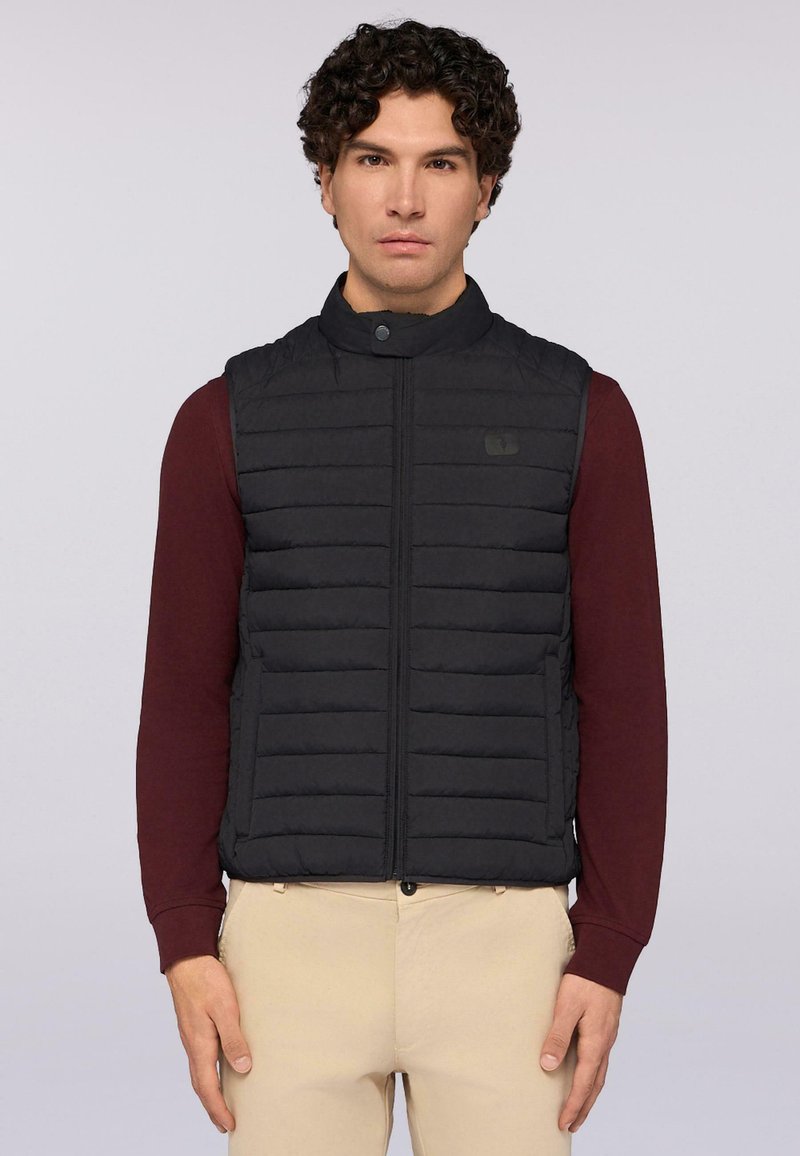 Černá vyp padded vesta s vysokým límcem, vertikálním prošíváním, předním zipem a logem. Nositá přes burgundské tričko s dlouhým rukávem.