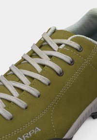 Κοντινή λήψη αθλητικού παπουτσιού σουέντ σε ελαιί πράσινο χρώμα με λευκά κορδόνια, ορατή ραφή και λογότυπο στο πλάι σε ανοιχτή σόλα.