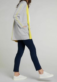 Ljusgrå cardigan med gula accenter, två framfickor och en avslappnad passform, parat med mörkblå jeans och vita sneakers.