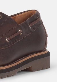 Brown leather shoe with a rounded toe, stitched detail, metallic eyelets, and a thick rubber sole featuring textured treads.