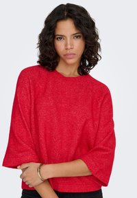 Pull rouge texturé à coupe décontractée, manches larges et encolure arrondie. Porté avec un bas noir et un bracelet doré au poignet.