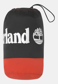 Sort og rød vandtæt pose med snor. Har et hvidt "Timberland" logo og et træsymbol, glat overflade, cylindrisk form.