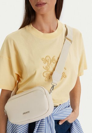 Mujer con una camiseta bordada de color amarillo pálido, una camisa azul a rayas anudada a la cintura y un bolso cruzado de color crema con una correa ancha.