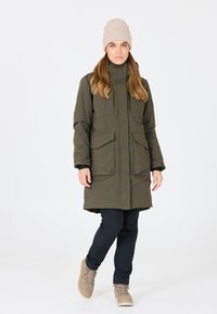 Olivgrön parkajacka med två stora framfickor, hög krage och dragkedja. Bärs över svarta byxor och bruna snörkängor.