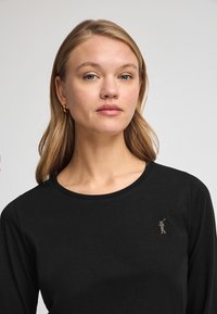 Svart långärmad tröja med rund halsringning som har ett litet, ljusbrunt emblem på vänster bröstområde. Mjuk bomullstyg.