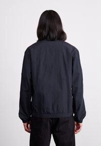 Chaqueta azul oscura hecha de material ligero. Presenta un ajuste relajado, puños elásticos y sutiles acentos rojos en los hombros.