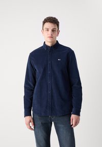 Camicia in velluto a coste di colore blu navy con colletto button-down, chiusura frontale con bottoni e un piccolo logo sul lato sinistro del petto. Tessuto testurizzato.