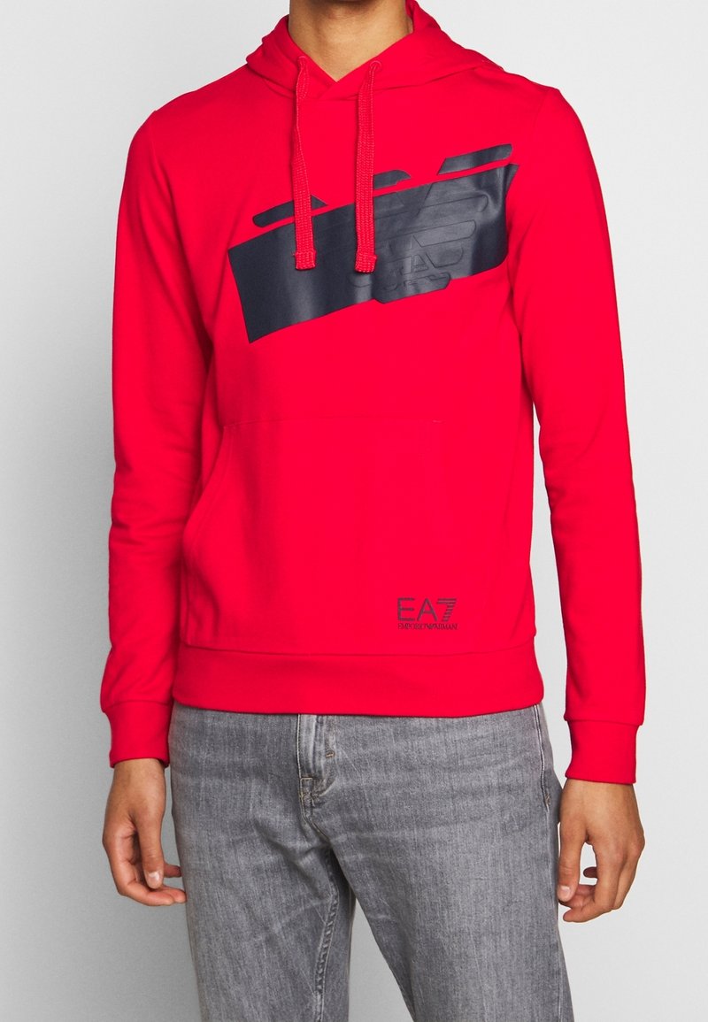 Czerwona bluza z kapturem wykonana z miękkiego materiału, z grafiką logo w ciemnoniebieskim kolorze na przodzie. Posiada przednią kieszeń oraz regulowane sznureczki kaptura.