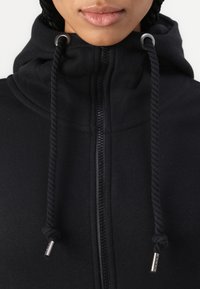 Veste à zip noire avec un col haut, des cordons de serrage avec des accents métalliques, et un tissu texturé pour la chaleur et le confort.