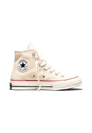 Beżowy, wysokogatunkowy sneaker Converse All Star z białymi sznurowadłami, czerwoną paskiem na podeszwie i okrągłym logo na kostce.