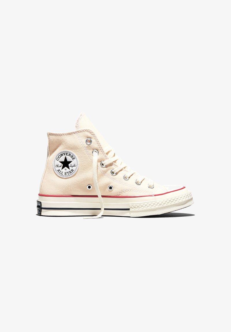 Baskets montantes beiges Converse All Star avec lacets blancs, bande rouge sur la semelle et logo circulaire sur la cheville.