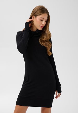 Jeune femme aux longs cheveux ondulés portant une robe pull à col roulé noire et de grandes boucles d'oreilles dorées en créoles sur un fond uni.