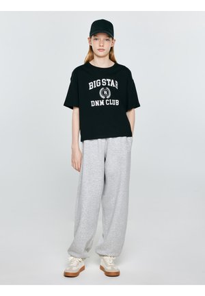 Młoda osoba ubrana w czarną czapkę, czarny t-shirt z napisem "Big Star DNM Club", szare spodnie dresowe i kremowe trampki, stojąca na jednolitym tle.