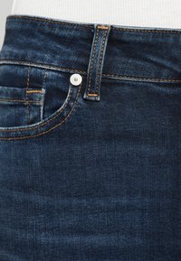 Blugii din denim albastru închis prezintă un design clasic cu cinci buzunare, cusături portocalii contrastante și o închidere cu nasture metalic la talie.