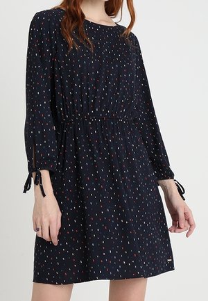 Mujer con vestido azul marino de mangas largas, cintura entallada y patrón de pequeños puntos rectangulares multicolor.