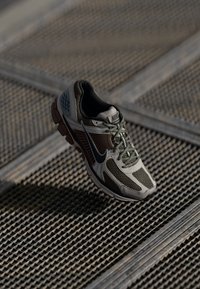 Nike futócipő, bézs hálós felső résszel, sötétszürke részletekkel, zöldes fűzőkkel és texturált talppal, egy rácsos felület fölött felfüggesztve.