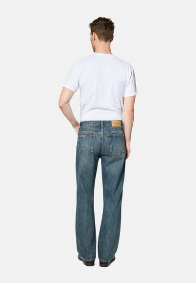 Man standing with hands on hips, wearing white short-sleeve t-shirt and blue straight-leg jeans, facing away against a plain background.