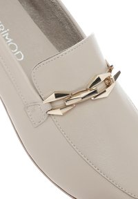 Scarpe slip-on in pelle beige con finitura texturizzata, caratterizzate da un accento di catena dorata sulla parte superiore e una punta rotonda e liscia.