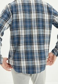 Camicia di flanella a quadri blu e bianca con dettagli neri, caratterizzata da un orlo a coda e polsini con bottoni. La texture appare morbida e tessuta.