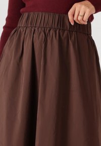 Bruine, vloeiende rok met een elastische tailleband; de stof lijkt licht en glad, met een subtiele glans en geplooide details.