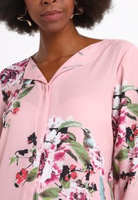 Blouse rose ornée d'un motif floral aux couleurs vives, en tissu léger, avec un col en V agrémenté d'un bouton.