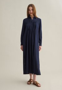 Abito camicia blu navy con maniche lunghe, chiusura frontale con bottoni, dettagli plissettati e spacchi laterali, realizzato in tessuto leggero, abbinato a sandali marroni.
