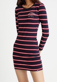 Mörk blå klänning med röda och vita horisontella ränder. Långa ärmar, rund halsringning och liten "Superdry"-logotyp på bröstet.