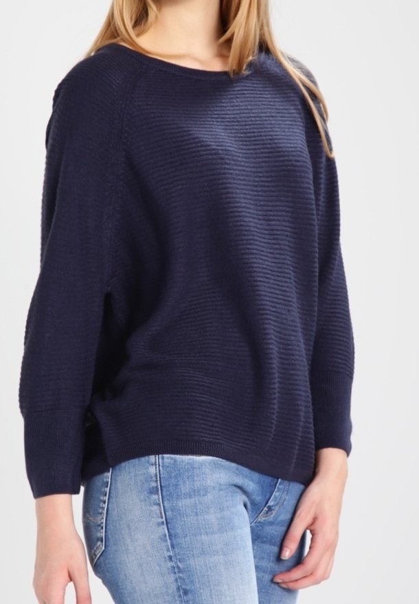 Femme portant un pull côtelé bleu marine à manches trois-quarts et un jean bleu clair sur un fond uni.