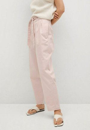 Osoba ubrana w jasnoróżowe, wysokie spodnie z tkaninowym paskiem, kremową bezrękawnikową dzianinową bluzkę i białe sandały typu slide, stojąca na jednolitym tle.