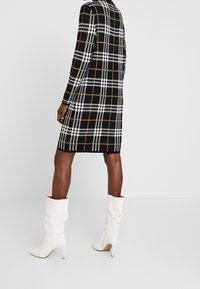 Personne portant une robe à carreaux noire, blanche et orange avec des manches longues et des bottes blanches à talons hauts et souples sur un fond uni.