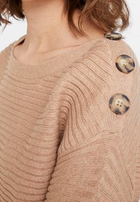 Beige ribstrikket sweater med en bred halsudskæring og tre dekorative mørke knapper på venstre skulder. Blød tekstur.
