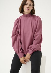 Blouse mauve avec col haut, surface texturée, manches bouffantes et poignets ajustés. Portée avec un pantalon noir, sur fond uni.