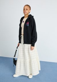 Svart hettejakke med fargerik grafikk, beige tettsittende topp, hvit lagdelt skjørt og kremfargede slip-on sko, som bærer en svart crossover-veske.