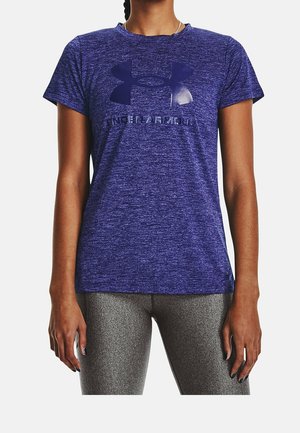 Vrouw draagt een paarse Under Armour T-shirt met korte mouwen en grijze legging, staat met ontspannen armen langs haar zij.