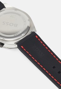 Correa de reloj de goma negra con superficie texturizada, costura roja y fondo de acero inoxidable grabado con "BOSS" y detalles de resistencia al agua.